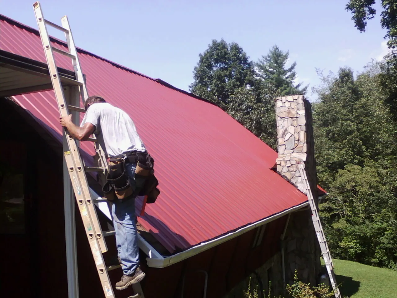 Roofer on a ladder beside a red metal roof and stone chimney — Soffit & Fascia Repair by Roofing Brothers in Sheboygan Falls
